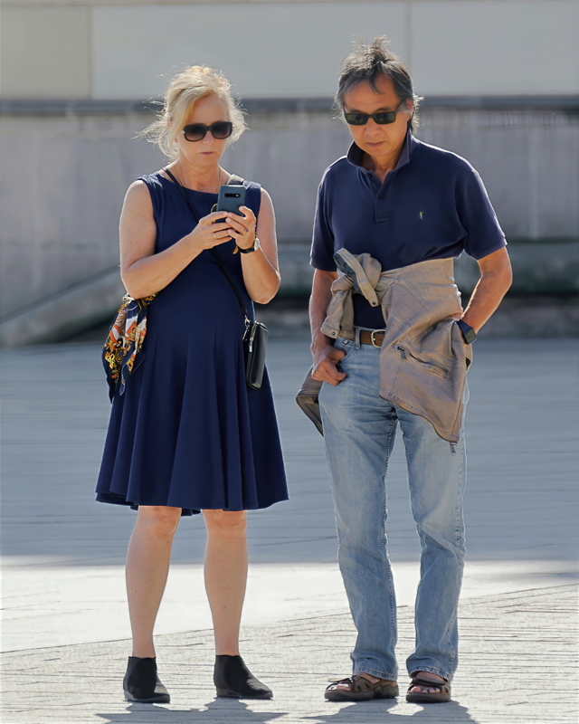 20 - Two individuals outdoors in casual attire, with the woman on the left looking at her mobile phone and the man on the right standing next to her wearing sunglasses and a polo shirt